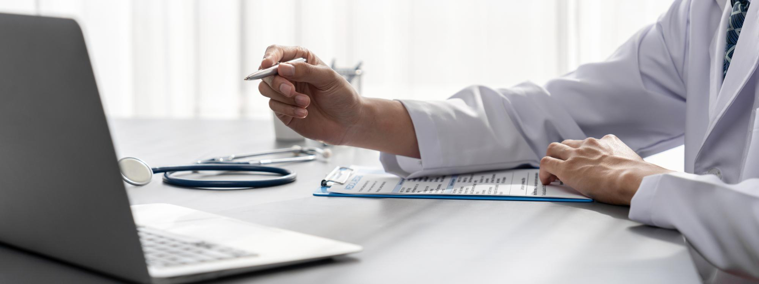 Κοντινό πλάνο στο χέρι ενός γιατρού που κρατάει ένα στυλό, με ένα ανοιχτό λάπτοπ στο γραφείο μπροστά του.